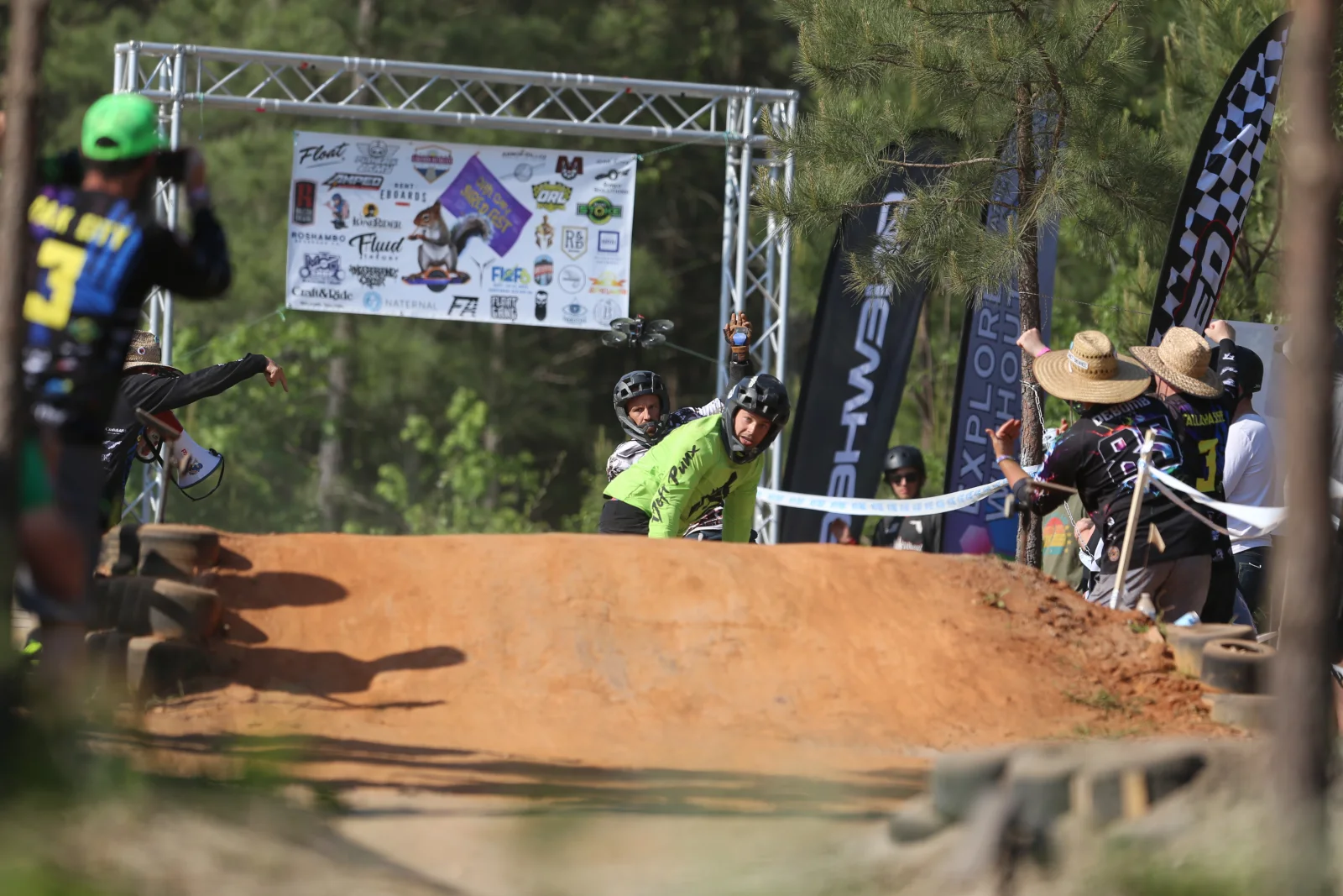 Onewheel racers cresting a dirt-course feature with spectators beside the course.